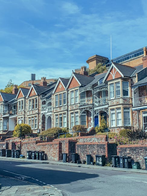 Image of a row of Victorian terraced houses with ornate white balconies and large bay windows, situated on a street with a brick wall and a line of black wheelie bins along the pavement. The houses have pitched roofs with decorative gables and are set against a bright blue sky with some wispy clouds. The scene represents a typical residential area suitable for home relocation or furniture transport, with the street appearing quiet and well-maintained, ideal for house removal services in Cricklewood. Man with Van Cricklewood offers professional removals and moving logistics within this neighbourhood, supporting packing, loading, and transport processes. The image captures the external environment before the moving process begins, with no visible activity or equipment currently in use.