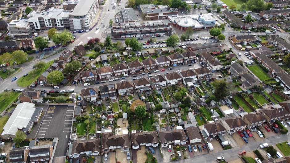 A street scene in Cricklewood showing several parked cars along the roadside and a few driving vehicles on the road, with trees lining the pavement and a historic church building with a clock tower and ornate architectural details in the background under a partly cloudy sky. The image captures a daytime view of the urban environment, where home relocation and furniture transport services by Man with Van Cricklewood may be coordinated, with the church and surrounding buildings providing context for local removals and moving logistics.