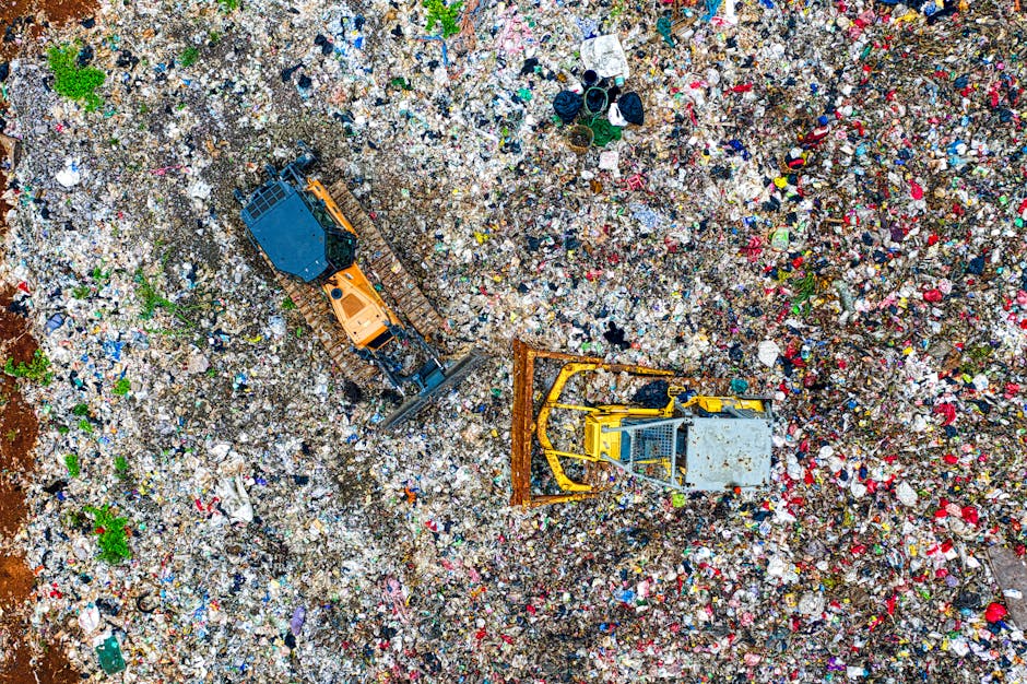 An aerial view of a large landfill site filled with mixed household waste, including plastic bottles, cardboard, fabric, and other debris scattered across the ground. Two large construction vehicles are actively engaged in the waste management process: a yellow bulldozer pushing and compacting the rubbish, and an orange excavator with a black arm and bucket lifting or moving materials. The scene appears to be outdoors under natural daylight, with the vehicles positioned amid the densely packed waste. This image reflects aspects of waste disposal and material handling relevant to relocation and moving services, highlighting the importance of proper waste management after a house move. Man with Van Cricklewood occasionally encounters such scenarios when assisting clients with post-move clearance and disposal, supporting the logistics of home relocation and packing.
