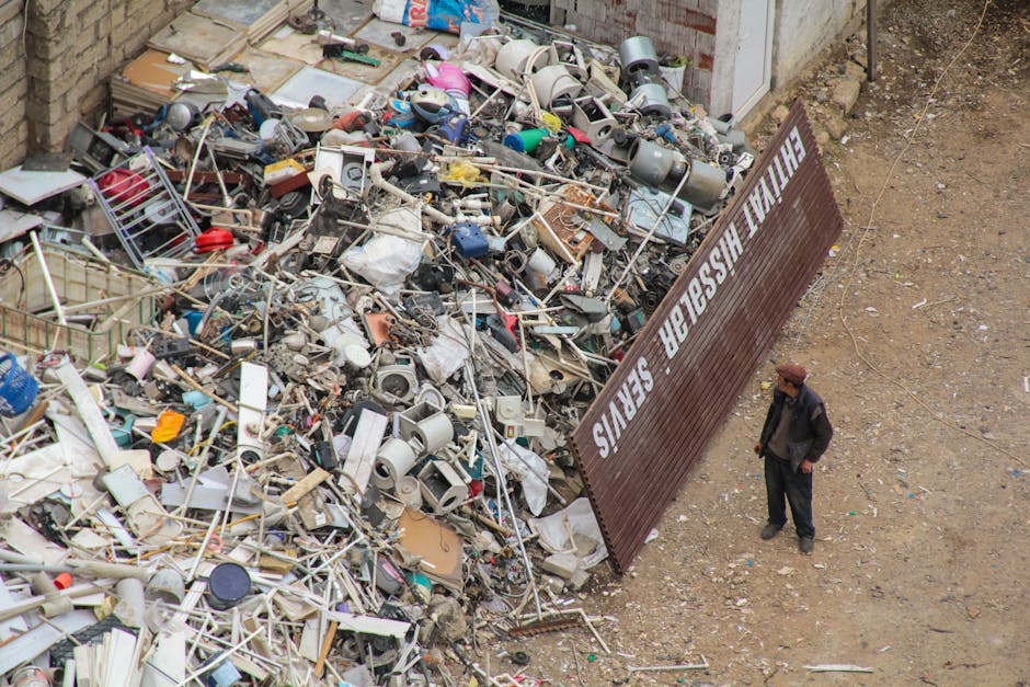 A large pile of assorted household waste, including broken appliances such as microwaves and small kitchen equipment, cardboard boxes, plastic containers, metal frames, and discarded electronics, is situated outdoors on a gravel surface next to a building wall. A man wearing a dark jacket, jeans, and a brown hat stands nearby, observing the site. A metal sign with the text 'HELVETIA FURNISHER SERVICES' leans against the pile, indicating possible waste disposal or charity collection. The scene appears to be part of an outdoor furniture and bulky item disposal process, with the waste materials awaiting removal or recycling as part of a home relocation or rubbish clearance service provided by Man with Van Cricklewood.