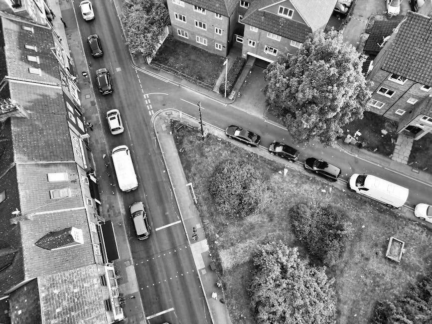 An aerial black-and-white photograph captures a narrow residential street during a house removal process, with several parked cars along the curb and on the street, including a large white van at the far end. The street is lined with closely spaced terraced houses and some detached homes, with front gardens and trees visible. In the vicinity, a small grassy area is bordered by a low fence, with a few walking paths and mature trees providing shade. The loading process appears to involve moving boxes and furniture, which are not visible in this outdoor scene. The vehicles are positioned to facilitate the packing and transport of household items as part of a home relocation service by Man with Van Cricklewood, working within the constraints of the narrow street and limited parking area.