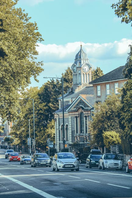 A view of a residential street during daytime with several cars parked along the curb on either side of the road. The street is lined with mature trees, some of which partially obscure a historic church building with a clock tower and a pointed spire in the background. The church features arched windows and a decorative stone façade, indicating traditional architecture. The sky is partly cloudy with patches of blue visible. The scene does not show any furniture, boxes, or moving activity, but this setting relates to house removals and relocation logistics, as indicated by the page titled 'Claremont Road Removals: Narrow Street Moving Guide' on manwithvancricklewood.co.uk.