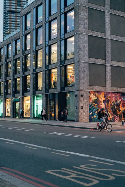 The image shows a multi-storey commercial building with a modern brick and glass façade, situated along a busy city street. The ground floor features large glass shopfronts with illuminated displays,