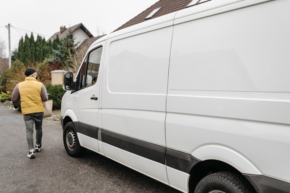 A man wearing a black cap, brown sleeves, and a yellow padded vest is walking on a residential street towards a large white commercial van with sliding side doors, parked outside a house with a sloped tiled roof and brick chimney. The van, which is part of a home relocation or furniture transport process, is positioned with its front facing left, close to the curb, with the driver's side mirror visible. The street features a mix of concrete pavement and residential landscaping including trees, bushes, and a small brick structure. In the background, there are neighboring houses with pitched roofs, electrical poles with wires, and cloudy sky overhead, indicating an overcast day. The scene depicts loading or unloading activity typical of professional removals, supported by the presence of packing materials or boxes inside the van or nearby, although not directly visible in this particular image. Man with Van Cricklewood is involved in this furniture transport or packing and moving process.