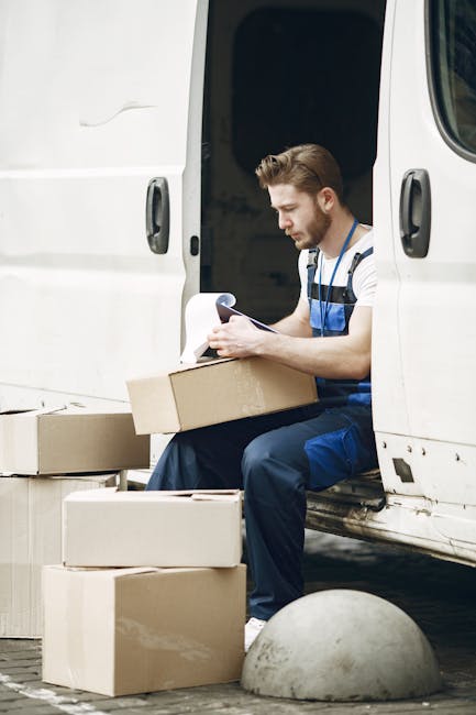 A male mover with light skin and short brown hair, wearing a striped t-shirt and blue work overalls, seated on the edge of an opened white van's doorway during a house removal. He is holding a clipboard and reviewing documents, with several cardboard boxes placed on the ground beside him and inside the van. The scene is set outdoors on pavement, evidencing a loading process involved in furniture transport and packing for a home relocation. The van's interior is visible behind him, and the environment appears well-lit, capturing the logistical aspect of moving services offered by Man with Van Cricklewood in the context of planning and executing house removals near Cricklewood Broadway.