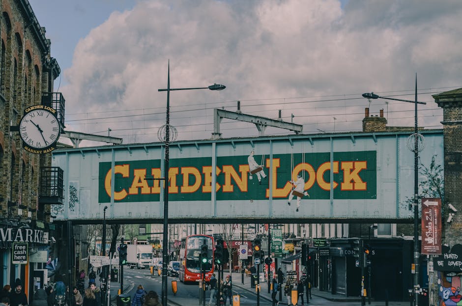 A busy street scene in Camden Lock featuring a large overhead railway sign painted with the words 'Camden Lock' in bold yellow and orange letters. The sign is mounted on a steel structure above the road, which is lined with various shops, including a market and retail outlets. Pedestrians are walking along the pavement, some carrying shopping bags, while others wait at traffic lights. A double-decker red bus is visible in the background, indicating public transport accessibility. Streetlights and overhead wires run across the scene, with a clock mounted on a building to the left, showing the time as approximately 12:20. The sky is partly cloudy, casting diffuse lighting over the area. This urban setting illustrates a typical street involved in home relocation or furniture transport, with various objects, including storefronts and signage, highlighting the busy, vibrant environment of Camden during the day. Man with Van Cricklewood occasionally operates in such busy locations, supporting efficient packing and moving logistics in the area.