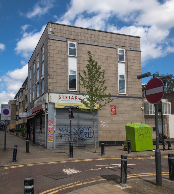 Exterior view of a three-story building with a beige brick facade and multiple vertical windows, situated on a street corner under a partly cloudy sky. The ground level features a closed storefront with a metal roller shutter covered in graffiti and posters, indicating a retail shop or service unit. A small tree with green leaves is planted in front of the building, slightly obscuring part of the storefront. To the right, a bright green waste or recycling bin with graffiti is placed near the sidewalk, alongside a black bollard barrier. A red and white 'no entry' road sign is mounted on a pole close to the building, with additional street signage visible further along the pavement. The street has yellow double lines indicating parking restrictions, and black bollards are positioned along the sidewalk to prevent vehicle access. The scene captures a typical urban environment, relevant to home relocation or furniture transport logistics, with the building possibly serving as a point of origin or destination in a moving process. Man with Van Cricklewood offers removals services in this area, supporting packing, loading, and transportation of household furniture and boxes.