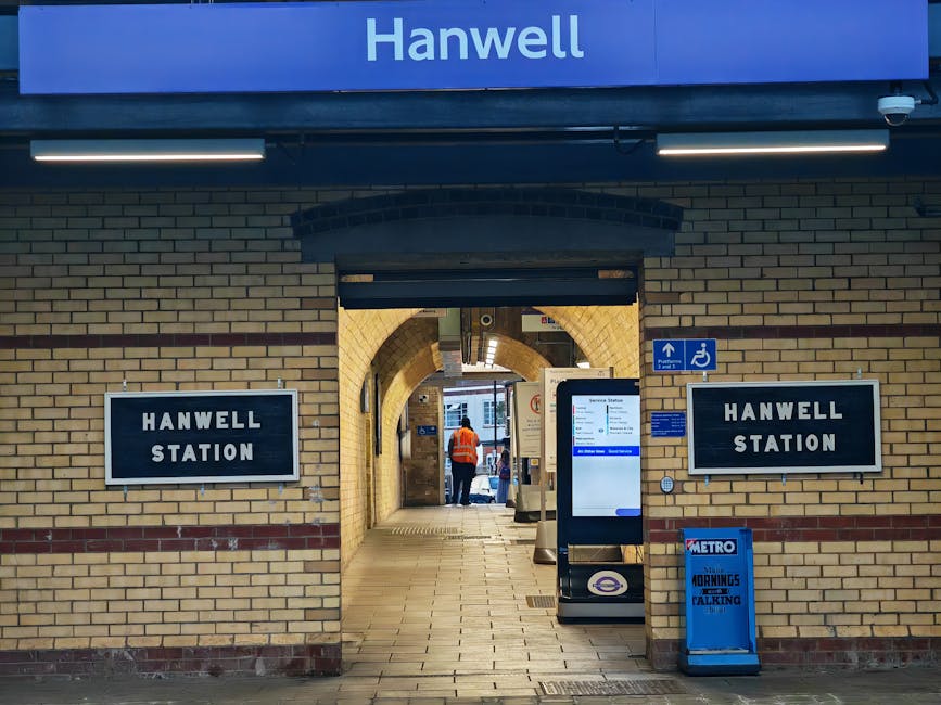 Inside the entrance of Hanwell Station, a brick-walled corridor leads to the platform area, with a digital information screen positioned on the right side near the doorway. Two black and white signs displaying 'HANWELL STATION' are mounted on the brick walls on either side of the entrance. A person wearing an orange high-visibility vest stands at the far end of the corridor, near the station's platform, which is visible through an arched brick doorway. Outside, the platform is illuminated by natural light, and part of a vehicle can be seen parked on the street. For a home relocation or furniture transport project, [COMPANY_NAME] can assist with packing and moving services, ensuring items such as boxes, furniture, and appliances are efficiently loaded and transported, as evidenced by the orderly scene at the station entrance.