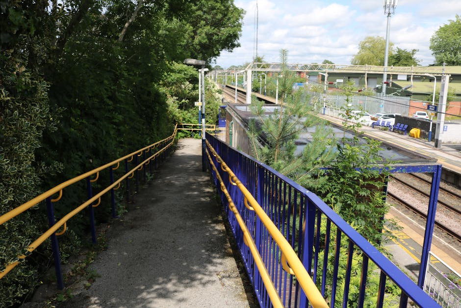 A narrow outdoor pedestrian walkway adjacent to a railway station, with a concrete surface and a blue metal railing on the right side, painted in yellow on top, guiding towards a staircase descend. To the left, dense green foliage and trees cast shadows across the pathway. In the background, railway tracks, platforms, and overhead electrical lines are visible, alongside parked cars and station infrastructure. The scene is illuminated by natural daylight, with partly cloudy skies overhead. This setting depicts an accessible route next to a train station, relevant for home relocation or furniture transport logistics involving the station area, and is associated with house removals and moving services as provided by Man with Van Cricklewood.
