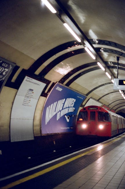 Inside the entrance of Hanwell Station, a brick-walled corridor leads to the platform area, with a digital information screen positioned on the right side near the doorway. Two black and white signs displaying 'HANWELL STATION' are mounted on the brick walls on either side of the entrance. A person wearing an orange high-visibility vest stands at the far end of the corridor, near the station's platform, which is visible through an arched brick doorway. Outside, the platform is illuminated by natural light, and part of a vehicle can be seen parked on the street. For a home relocation or furniture transport project, [COMPANY_NAME] can assist with packing and moving services, ensuring items such as boxes, furniture, and appliances are efficiently loaded and transported, as evidenced by the orderly scene at the station entrance.