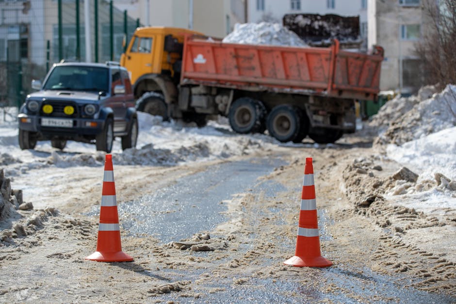 A construction or moving site on a snow-covered residential street with orange and white traffic cones placed on a dirt and snow mixture to block access. In the background, there is a yellow dump truc