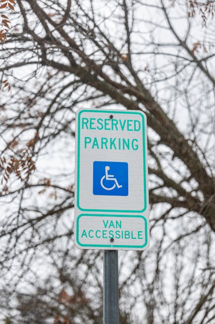 A reserved parking sign indicating accessible parking for disabled individuals and van accessibility, mounted on a metal pole against a background of leafless tree branches and a cloudy sky. The sign features a blue and white wheelchair symbol and text stating 'RESERVED PARKING' and 'VAN ACCESSIBLE.' This signage is positioned near a property entrance, relevant to house removals and home relocation services, with the nearby environment suggesting an urban or suburban area. Such parking permits are essential for smooth furniture transport and logistics during moving processes, as managed by companies like Man with Van Cricklewood, supporting efficient access for loading and unloading items inside the property.