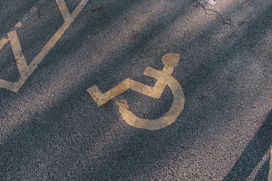 A close-up view of a designated accessible parking space on an asphalt surface, featuring a yellow paint symbol of a person using a wheelchair. The symbol is worn and partially faded, with visible cracks and texture in the asphalt. Nearby, part of another parking space marking can be seen, indicating organized parking guidelines. The area appears to be outside a residential or commercial property, with natural light illuminating the surface. This image reflects parking arrangements that facilitate home relocation and furniture transport, especially for individuals with mobility needs, relevant to house removals and moving services provided by Man with Van Cricklewood, as referenced on the webpage titled 'Gladstone Park Removals: Access, Parking and Best Routes, CRICKLEWOOD'.