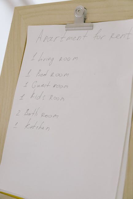 A handwritten list on a white sheet of paper attached to a wooden clipboard with a metal clip, detailing rooms in an apartment for tenancy clearance. The list includes 'Living room,' 'Bedroom,' 'Guest room,' 'Kids room,' 'Bathroom,' and 'Kitchen,' written in pencil. The clipboard appears to be placed on a wooden surface inside a property, likely used for organizing packing or moving tasks related to a house removal or relocation services. The background shows part of a wall with neutral lighting, appropriate for documenting the contents or planning of the move, consistent with a professional furniture transport and packing process facilitated by Man with Van Cricklewood.