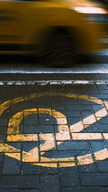 The image shows a section of a paved street with a yellow wheelchair parking symbol painted on the dark, textured bricks. A yellow van is captured in motion above the parking space, with part of its body and wheel visible, slightly blurred due to movement. The van is parked adjacent to the designated parking area, which is marked clearly on the pavement to indicate accessible parking for individuals with disabilities. In the background, the environment suggests an urban setting, potentially near a residential or commercial building. The scene relates to parking regulations in Cricklewood, relevant to house removals and moving services provided by Man with Van Cricklewood, highlighting the importance of understanding parking rules during furniture transport and home relocation processes.