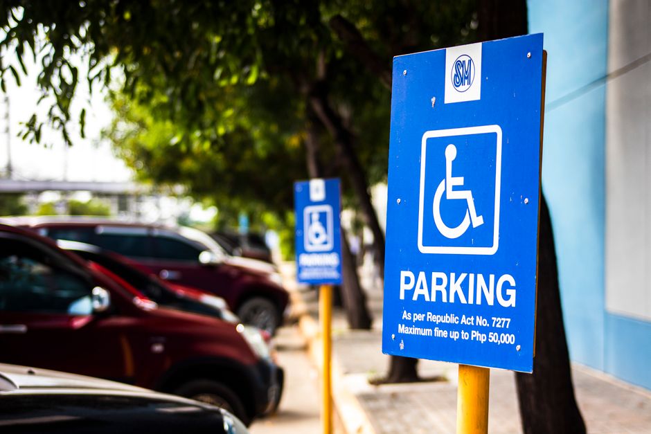 A bright blue parking sign indicating designated parking for disabled individuals, with white text stating 'PARKING' and referencing the Republic Act No. 7277, along with a white wheelchair symbol. The sign is mounted on a yellow pole located on a paved pavement area next to parked cars, including a black vehicle and a maroon one, with more similar signage visible in the background. The scene is outdoors with a row of trees providing shade, and sunlight illuminating the area. This image captures a parking zone near a property, relevant to house removals and moving logistics, illustrating considerations for parking regulations during home relocation or furniture transport, with the sign referencing parking rules that may impact loading and unloading processes.