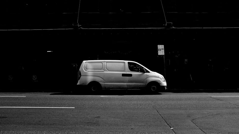 The image shows a section of a paved street with a yellow wheelchair parking symbol painted on the dark, textured bricks. A yellow van is captured in motion above the parking space, with part of its body and wheel visible, slightly blurred due to movement. The van is parked adjacent to the designated parking area, which is marked clearly on the pavement to indicate accessible parking for individuals with disabilities. In the background, the environment suggests an urban setting, potentially near a residential or commercial building. The scene relates to parking regulations in Cricklewood, relevant to house removals and moving services provided by Man with Van Cricklewood, highlighting the importance of understanding parking rules during furniture transport and home relocation processes.