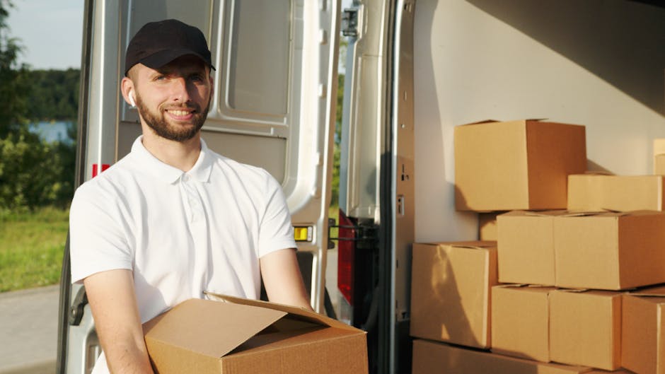A man standing on a residential street outside a house with a white gabled roof, loading medium-sized cardboard boxes into the open rear doors of a white van parked on the side of the road. The boxes